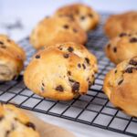 Nahaufnahme von sechs goldbraunen, veganen Schokobrötchen mit Schokostückchen auf einem schwarzen Gitter. Ideal für ein schokoladiges Frühstück.