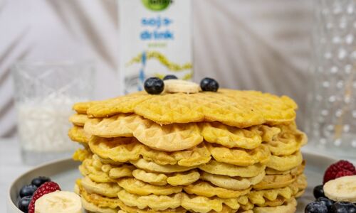 Ein Stapel frischer, goldbrauner veganer Waffeln auf einem grauen Teller, dekoriert mit Bananenscheiben und Blaubeeren. Im Hintergrund ein Sojadrink von Veganz.