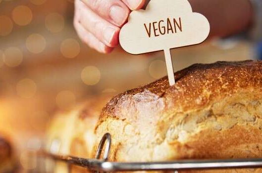 Nahaufnahme von frischem veganem Brot mit golden-brauner Kruste in einem Brotkorb, Hand hält veganen Holzschild.