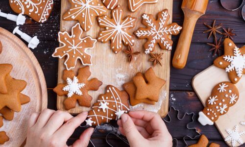 Eine Person dekoriert vegane Lebkuchenplätzchen mit Zuckerguss auf einem Holzbrett in einer weihnachtlichen Küche.