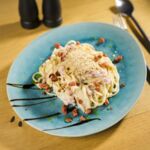 Vegane Carbonara auf blauem Teller mit veganem Parmesan und pflanzlichen Speckwürfeln, dekoriert mit Balsamico-Glasur.