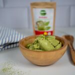 Grüne, vegane Matcha-Eiscreme in einer Holzschale, vor einer Tüte Matcha-Pulver im Hintergrund. Leckere vegane Alternative!