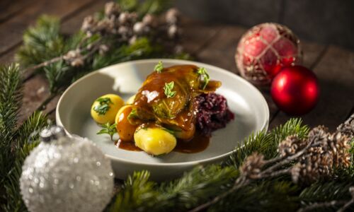 Ein festlich serviertes Gericht mit einer veganen Wirsingroulade in veganer Bratensoße, begleitet von kleinen Kartoffeln mit Kräutern und Rotkohl. Das Arrangement wird von weihnachtlicher Dekoration wie Tannenzweigen, Zapfen und roten Kugeln eingerahmt.