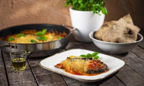 Vegane Parmigiana di Melanzane auf Teller vor Pfanne und Brot auf Holztisch, vegan und lecker.