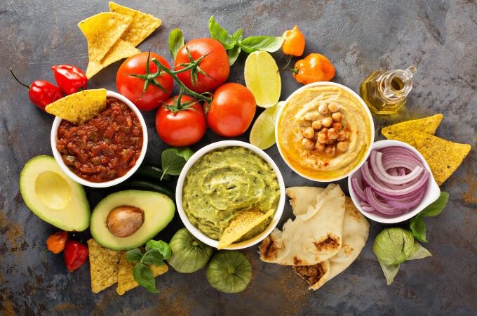 Bunte Auswahl an selleriefreien, veganen Lebensmitteln: Guacamole, Hummus, Salsa, frische Tomaten, Avocado, Limetten, Zwiebeln und Tortilla-Chips.