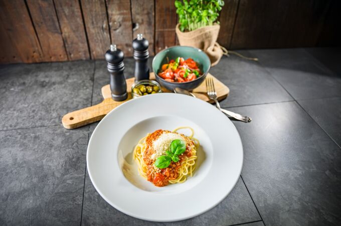 Ein stilvoll angerichteter Teller mit veganer Bolognese mit Spaghetti, garniert mit frischem Basilikum und veganem Parmesan. Im Hintergrund stehen Gewürzmühlen, ein kleiner Olivenbehälter und eine Schüssel mit frischem Tomatensalat.