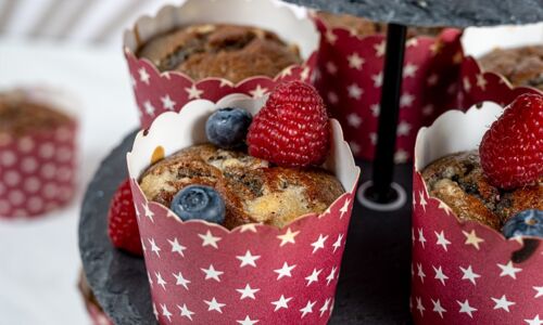 Vegane Beerenmuffins in roten Förmchen mit Sternenmuster, garniert mit Himbeeren und Blaubeeren auf einer schwarzen Etagere. Perfekt für vegane Naschkatzen!