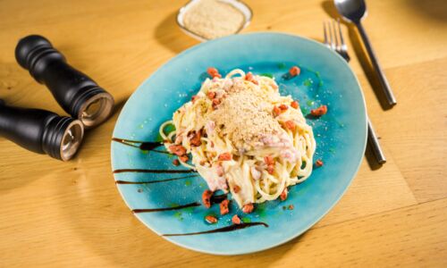 Vegane Carbonara mit Spaghetti, veganen Speckstückchen und veganem Parmesan auf blauem Teller, Pfeffermühlen im Hintergrund.