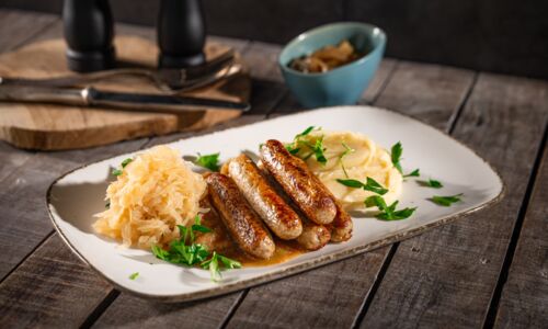 Vegane Würstchen (Mini-Griller) mit cremigem Kartoffelpüree, Sauerkraut und Schmorzwiebeln auf rustikalem Teller angerichtet.
