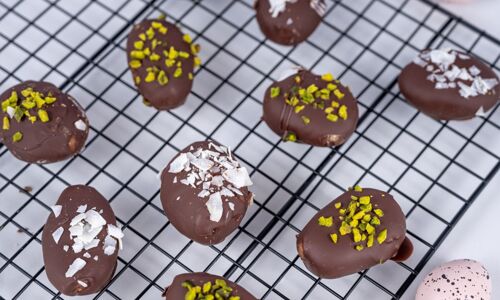 Acht vegane Erdnussbuttereier mit Schokoladenüberzug, Pistazien und Kokosflocken auf einem Gitter. Perfekt für Ostern.