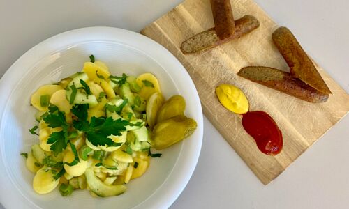 Vegane Kräutergriller auf Holzbrett mit Senf, Ketchup und Kartoffelsalat auf weißem Teller, dekoriert mit Petersilie.