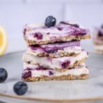 Nahaufnahme von veganen Blaubeer-Cheesecake-Bars, gestapelt auf einem Teller mit frischen Blaubeeren im Vordergrund.