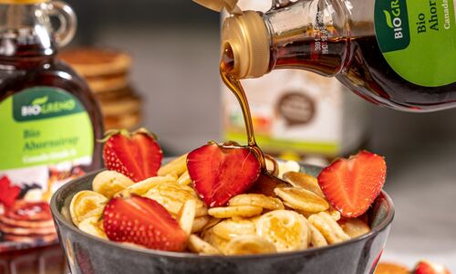 Eine Schale mit veganen Mini-Pancakes, dekoriert mit frischen Erdbeeren und übergossen mit Biogreno Bio Ahornsirup. Ideal für ein köstliches veganes Frühstück!