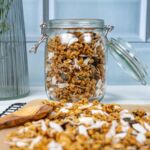 Glasbehälter mit knusprigem veganem Granola aus Haferflocken, Nüssen und Kokosflocken vor einem hellblauen Hintergrund, perfekt für ein gesundes Frühstück!