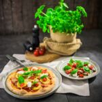 Frisch gebackene vegane Pizza mit gelber Zucchini, Tomaten, veganem Käseersatz und Basilikum, serviert auf einem glutenfreien Boden, daneben ein Teller mit Tomaten-Caprese-Salat und frischem Basilikum.