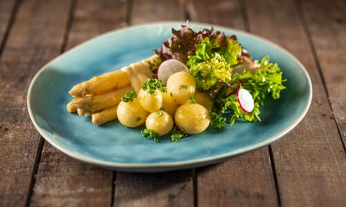 Nahaufnahme von weißem Spargel mit veganer Sauce Hollandaise, kleinen Kartoffeln und gemischtem Salat auf blauer Platte.