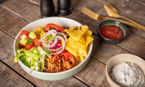 Vegane Burrito Bowl mit pflanzlicher Hack-Alternative, Salat, Tomaten, Gurken, Mais, roten Zwiebeln und Tortilla-Chips, serviert mit Salsa und cremigem Dip.