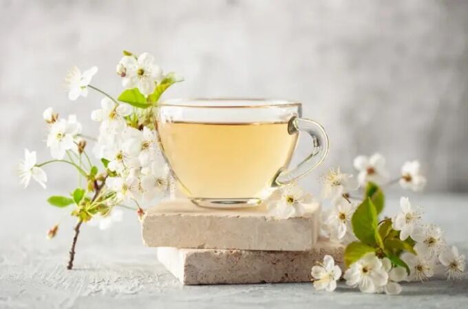 Gläserne Tasse mit hellem, veganem Tee, die auf zwei rechteckigen Steinplatten platziert ist. Um die Tasse herum sind zarte weiße Blüten mit grünen Blättern arrangiert.