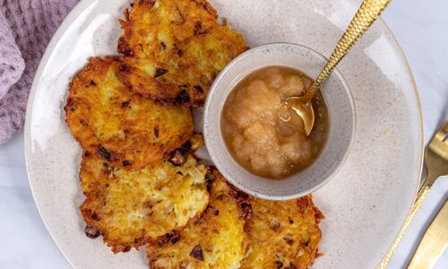 Teller mit vier goldbraunen veganen Kartoffelpuffern und Schale Apfelmus mit goldfarbenem Löffel, lilafarbenes Tuch im Hintergrund. Köstlich und vegan!