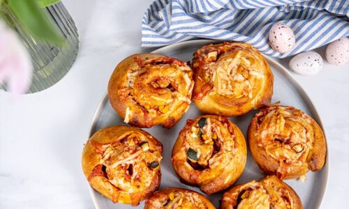 Sechs vegane, goldbraune Pizzaschnecken mit Gemüsestücken auf grauem Teller, flankiert von Tulpen und dekorativen Ostereiern. Ideal für österliche Anlässe!