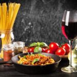 Vegane Spaghetti Bolognese in einer Pfanne neben Tomaten, Basilikum, Gewürzen und einem Glas Rotwein.