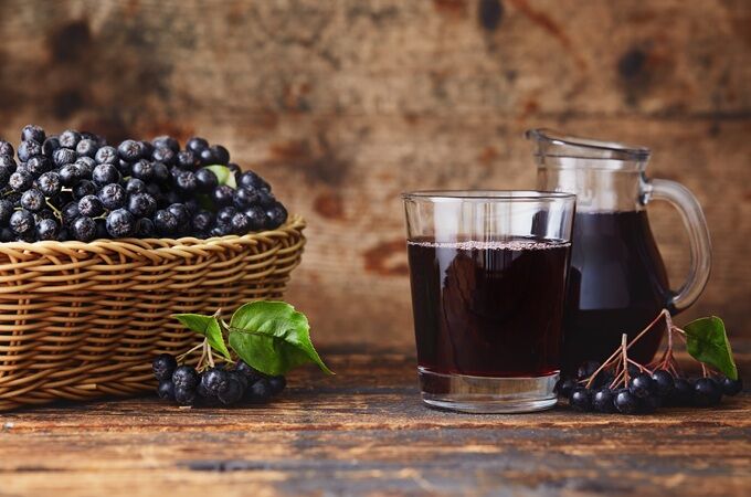 Ein Glas veganer Holundersaft neben einem Krug und einem Korb voller Holunderbeeren auf Holztisch.