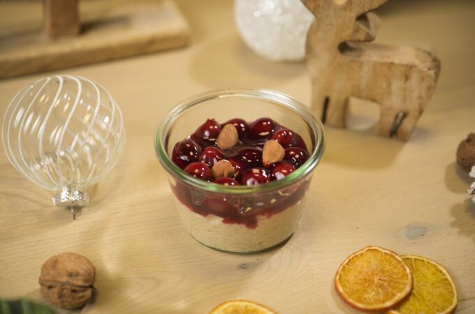 Ein Glas veganer Milchreis mit Kirschsoße und Mandeln auf einem weihnachtlich dekorierten Tisch mit Walnüssen und getrockneten Orangenscheiben.