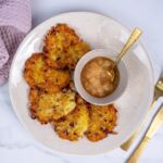 Teller mit vier goldbraunen veganen Kartoffelpuffern und Schale Apfelmus mit goldfarbenem Löffel, lilafarbenes Tuch im Hintergrund. Köstlich und vegan!