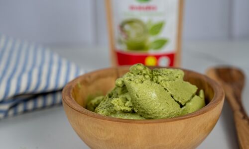 Grüne, vegane Matcha-Eiscreme in einer Holzschale, vor einer Tüte Matcha-Pulver im Hintergrund. Leckere vegane Alternative!