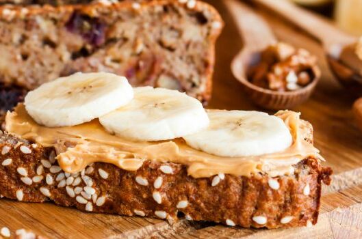 Eine Scheibe Körnerbrot mit veganem Erdnuss-Aufstrich und Bananenscheiben, im Hintergrund Nüsse und Aufstrich.