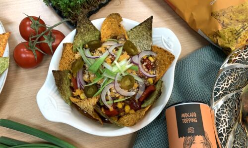 Bunte Schüssel veganer Tacoauflauf mit Mais, Jalapeños und roten Zwiebeln, umgeben von Tortilla-Chips. Frische Tomaten und Gewürze.