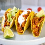 Drei knusprige vegane Tacos gefüllt mit Gemüse, veganem Fleisch und Soße auf einem Teller mit Limette im Vordergrund.