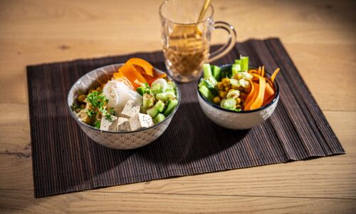 Zwei Schalen mit veganer Glasnudel-Bowl bestehend aus Glasnudeln, Tofu, Gemüse wie Gurken und Karotten auf einem braunen Tischset.