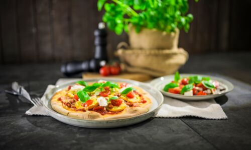 Vegane Pizza Frisch gebackene vegane Pizza mit buntem Gemüse, veganem Käseersatz und frischem Basilikum, serviert auf einem rustikalen Teller neben einem aromatischen Tomaten-Caprese-Salat