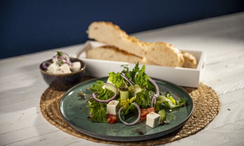 Veganer griechischer Salat auf grünem Teller mit roter Zwiebel, Tomaten, Gurke und veganem Käse. Im Hintergrund Brot.