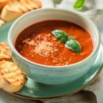 Vegane Tomatensuppe in einer grünen Schale, garniert mit Basilikum und veganem Parmesan, serviert mit geröstetem Brot. Probieren Sie den Genuss!