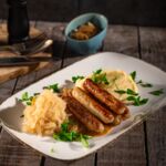 Gebratene vegane Würstchen mit Kartoffelpüree, Sauerkraut und Schmorzwiebeln, mit Petersilie garniert auf rustikalem Teller