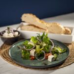 Veganer griechischer Salat mit veganem Feta, roten Zwiebeln, Gurken und Tomaten auf grünem Teller, serviert mit Brot im Hintergrund.