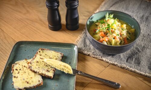 Warme, vegane Mahlzeit bestehend aus einer Schale mit Weißkohlsuppe und einem Teller mit zwei Scheiben Vollkornbrot, die mit pflanzlicher Margarine bestrichen sind. Die Suppe ist mit frischen Kräutern, Karotten und weiteren Gemüsestücken verfeinert.