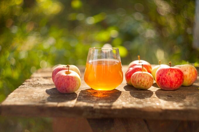 Ein Glas veganer Apfelsaft auf einem Holztisch, umgeben von frischen Äpfeln im Freien, unter sonnigem Licht.