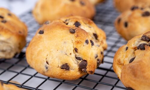 Nahaufnahme von sechs goldbraunen, veganen Schokobrötchen mit Schokostückchen auf einem schwarzen Gitter. Ideal für ein schokoladiges Frühstück.