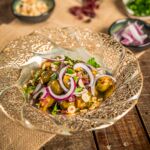 Geröstete Rosenkohlpfanne mit roten Zwiebelringen, gehackten Haselnüssen, Cranberrys und frischen Kräutern, serviert auf einem verzierten Teller auf rustikalem Holztisch mit kleinen Schälchen im Hintergrund.