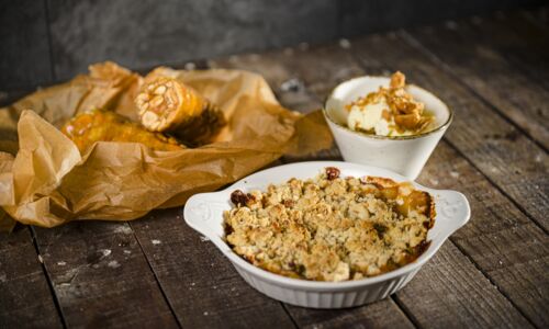 Apfel Crumble in weißer Keramikauflaufform mit Mandeln und Haferflocken auf rustikalem Holztisch, daneben ist ein Napf Vanilleeis mit Karamellsauce.