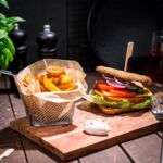 Vegane Crispy-Burger auf Holzbrett mit Gemüse, Salat und Pommes in Metallkorb, dekoriert mit Basilikum und Gewürzen, auf rustikalem Holztisch