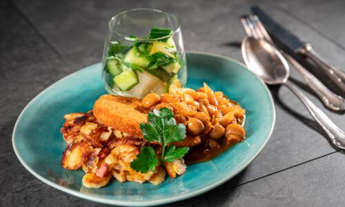 Gelbes veganes Schnitzel mit knusprigen Bratkartoffeln, Pilzsauce und grünem Salat in einem Glas auf blauem Teller.