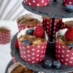 Vegane Beerenmuffins in roten Förmchen mit Sternenmuster, garniert mit Himbeeren und Blaubeeren auf einer schwarzen Etagere. Perfekt für vegane Naschkatzen!