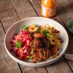 Bunte Bowl mit veganen Köfte, Couscoussalat, Rote-Bete-Weißkrautsalat, Granatapfelkernen und würziger Sriracha-Mayo auf rustikalem Holztisch.
