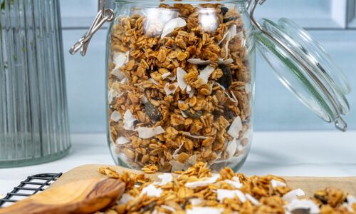 Glasbehälter mit knusprigem veganem Granola aus Haferflocken, Nüssen und Kokosflocken vor einem hellblauen Hintergrund, perfekt für ein gesundes Frühstück!