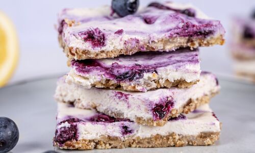 Nahaufnahme von veganen Blaubeer-Cheesecake-Bars, gestapelt auf einem Teller mit frischen Blaubeeren im Vordergrund.