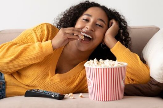 Frau liegt auf Sofa, isst veganes Popcorn aus roter Schüssel, lächelt und entspannt sich. Gemütlicher Abend mit veganen Snacks.